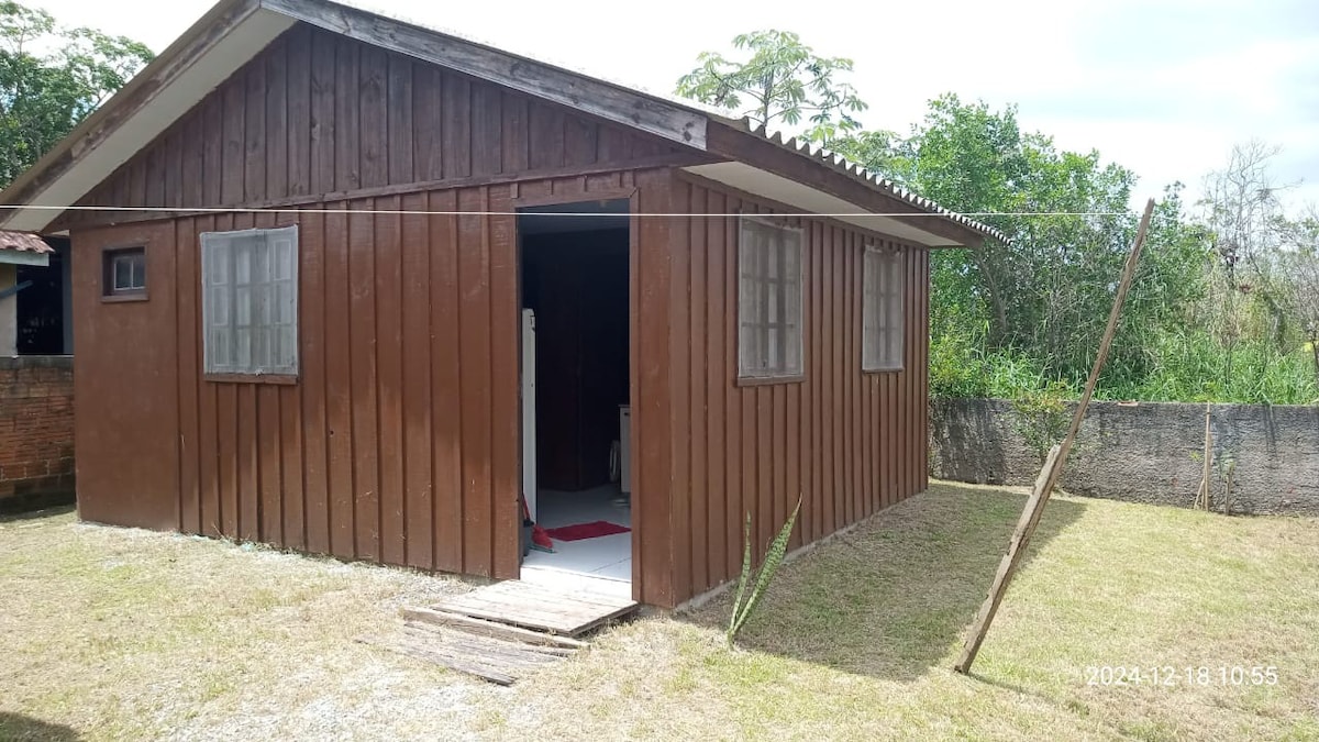 A simple wooden house with a brown exterior is visible, featuring two windows on the front. A partially opened door invites entry, revealing a glimpse of the interior. The surrounding area is grassy with scattered greenery, emphasizing a tranquil outdoor space.