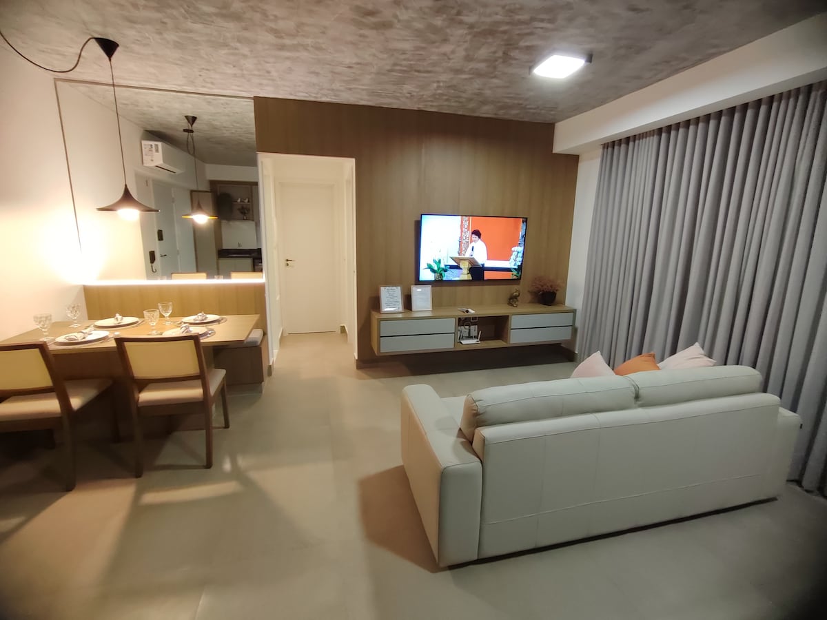 The living area features a modern design with a light-colored sofa facing a flat-screen TV mounted on the wall. A dining table with four chairs is positioned nearby, complemented by soft lighting for relaxation. Floor-to-ceiling curtains frame a window, allowing natural light to enter.