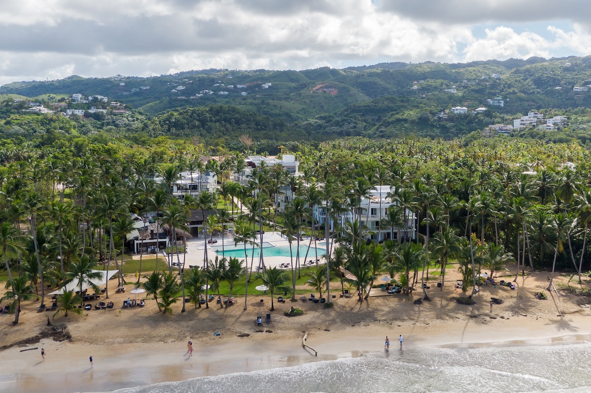 An aerial view showcases a lush landscape featuring a sandy beach lined with palm trees. A tranquil swimming pool is centrally located, surrounded by well-maintained gardens. The backdrop includes rolling hills and additional buildings, contributing to a serene coastal atmosphere.