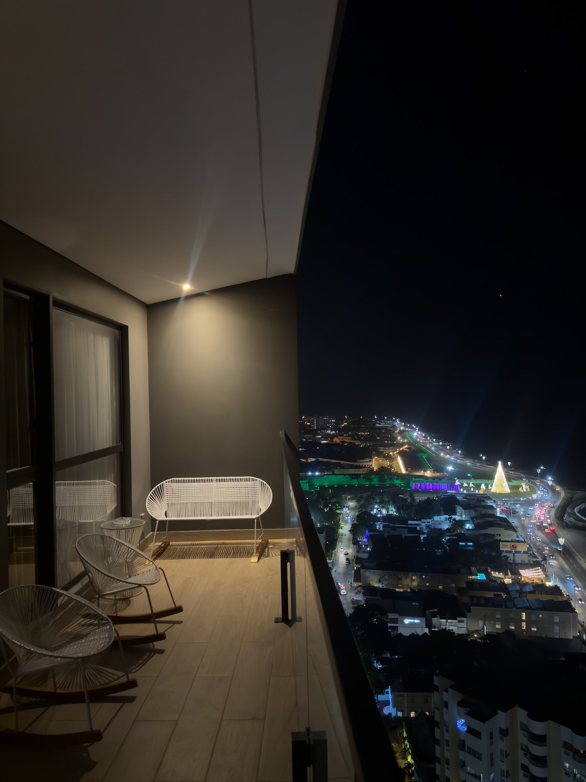 A balcony offers a view of the illuminated city at night, with twinkling lights visible in the distance. Two woven chairs and a small table are positioned on the wooden floor, while large glass doors provide access to the interior space.