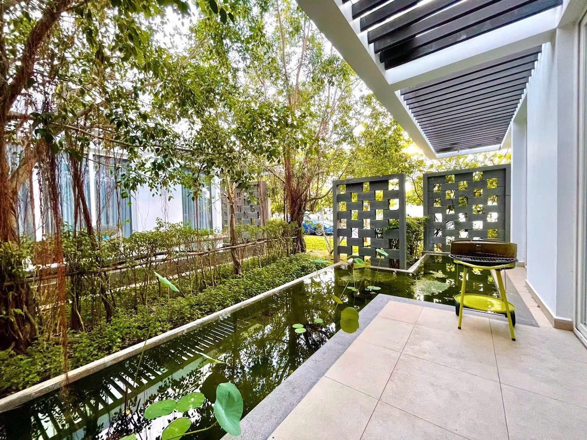 A serene outdoor area is surrounded by lush greenery, featuring a tranquil water feature with lily pads. A small table and chair are positioned nearby, providing a peaceful spot for relaxation. Natural light filters through the tree leaves, enhancing the calming atmosphere.