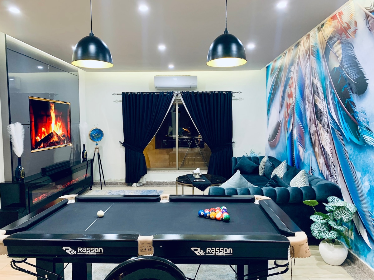 A spacious living area features a pool table at the center, surrounded by a modern dark sofa. An electric fireplace is integrated into the wall, and large windows with black curtains reveal a scenic view outside. Bold wall art adds a unique touch to the decor.