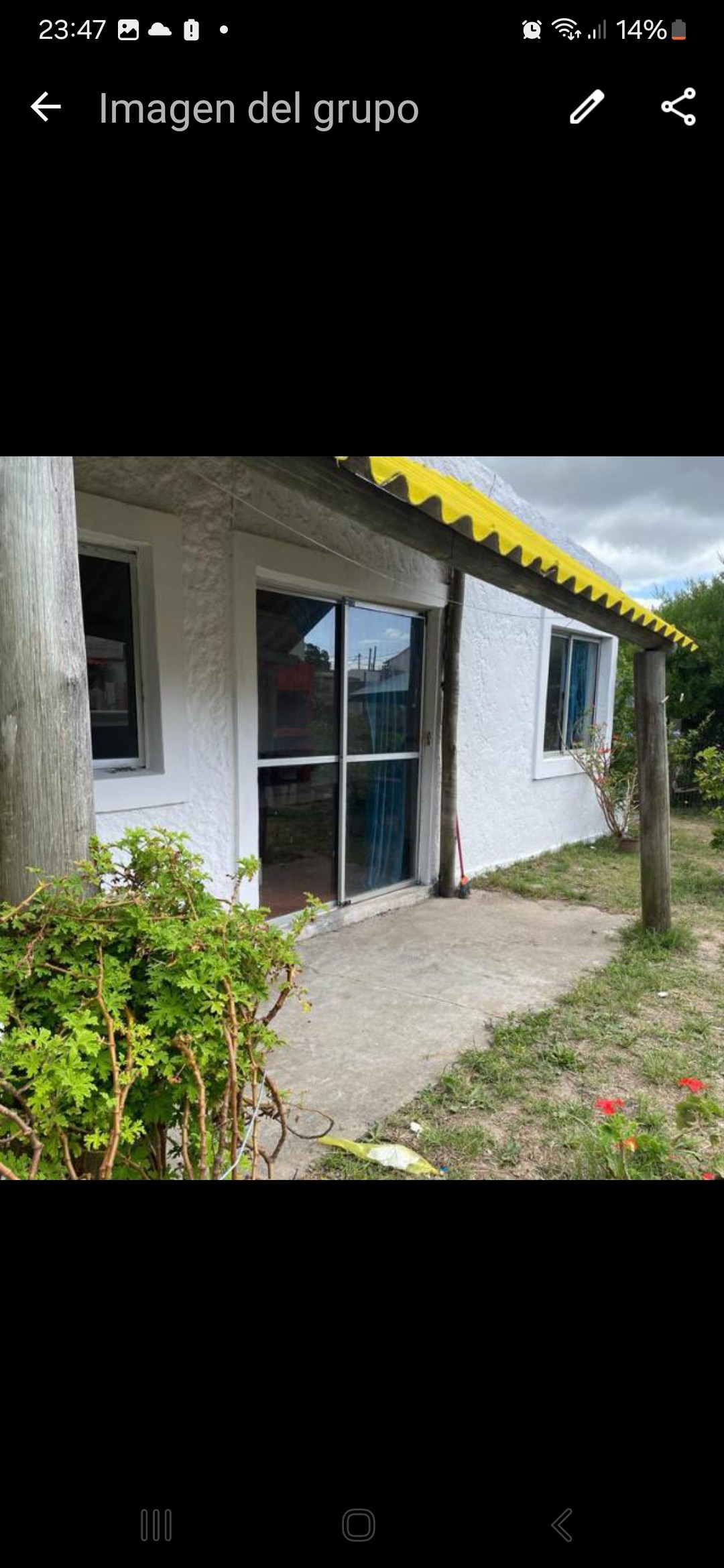 An exterior view of the house is presented, showcasing a white structure with large glass sliding doors that lead inside. A yellow canopy adds a pop of color above the entrance, while greenery and flowers are visible in the surrounding area.