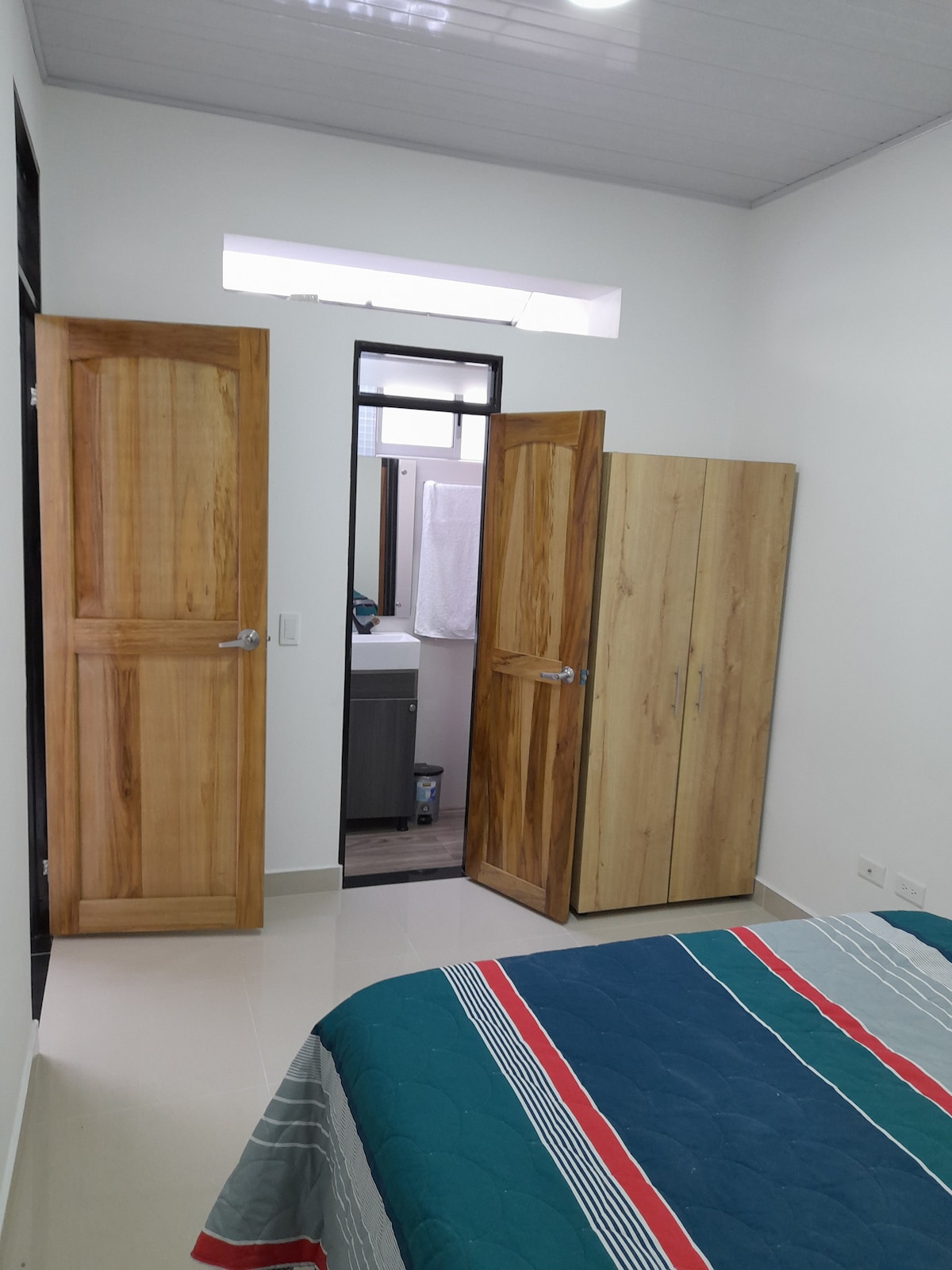 A well-lit bedroom features a spacious bed covered with a striped blanket. Two wooden doors lead to a bathroom and closet, while soft natural light enters through a small window above the bathroom entrance.
