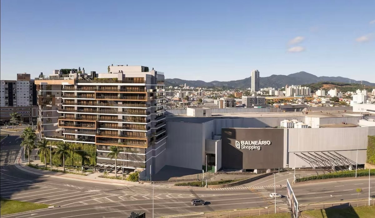 An aerial view captures the modern structure of Balneário Shopping, highlighted by its sleek design and surrounding landscape. The building features multiple balconies, with green spaces and palm trees lining the streets, complemented by distant mountains under a clear blue sky.