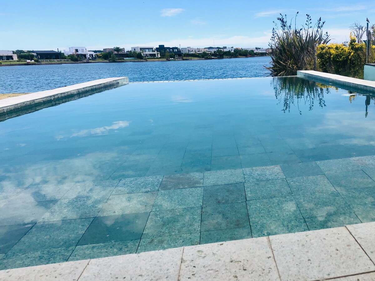 An infinity pool reflects the clear sky and the surrounding landscape. Its smooth, tiled edge seamlessly connects with the tranquil water of the nearby lagoon, which is framed by lush greenery and distant modern homes.