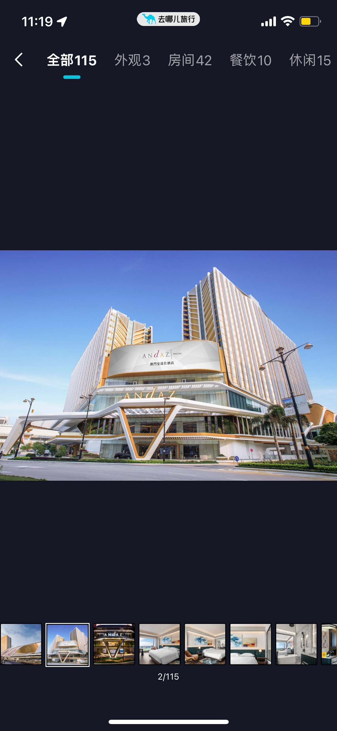 The exterior of a contemporary resort is captured, featuring modern architecture with large windows and a distinctive overhang. The building is situated in a well-lit urban area, framed by palm trees and a clear blue sky.