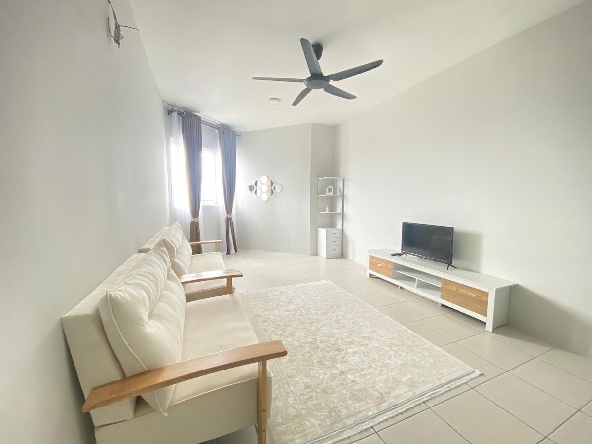 A spacious living area features a light-colored rug centered on tiled flooring. Two white sofas with wooden arms are positioned towards a small media unit and a television. Natural light enters from the window covered by sheer curtains, which are accompanied by dark blue drapes.