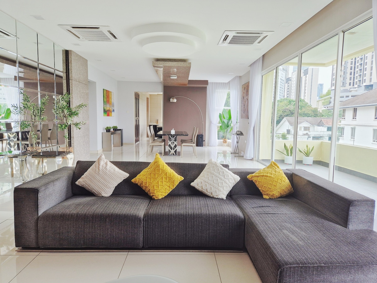 A spacious living area features a dark gray sectional sofa adorned with textured cushions. Large floor-to-ceiling windows allow natural light to fill the room, showcasing a view of greenery outside. A dining area with stylish chairs is visible through a glass partition.