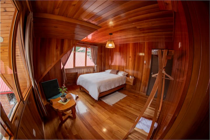 Habitación de madera con cama matrimonial, sofa, mesa de centro y colgadores para ropa.
Ubicada en el segundo nivel de la casa.