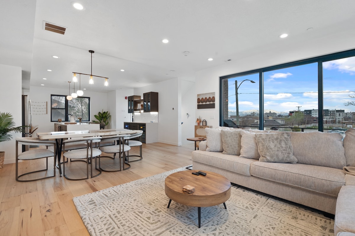 An open-concept living area is showcased, featuring a spacious sectional sofa facing a circular wooden coffee table. A dining area with a long table is visible, alongside a modern kitchen. Floor-to-ceiling windows provide natural light and views of the outdoors.