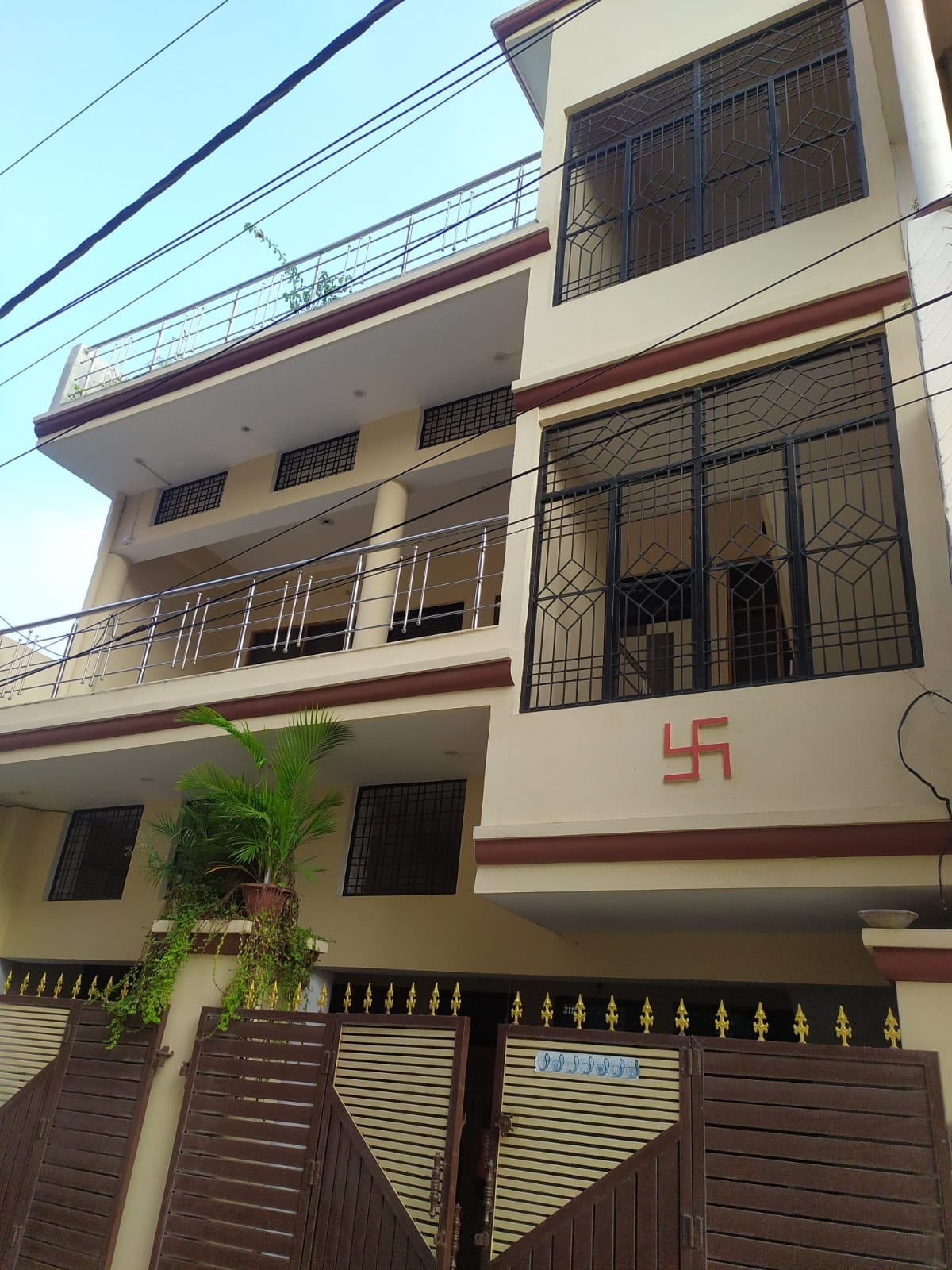The exterior of the building is shown, featuring a three-story structure with a balcony on each floor. A decorative plant is positioned at the entrance, and a wrought iron gate provides secure access. The façade is painted in neutral tones with geometric window designs.