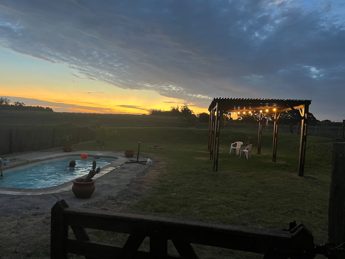 An outdoor area features a private pool, surrounded by green grass and a wooden pergola with string lights. The sunset casts warm hues across the sky, creating a serene ambiance. Two white chairs are positioned nearby, offering a spot to relax while enjoying nature.