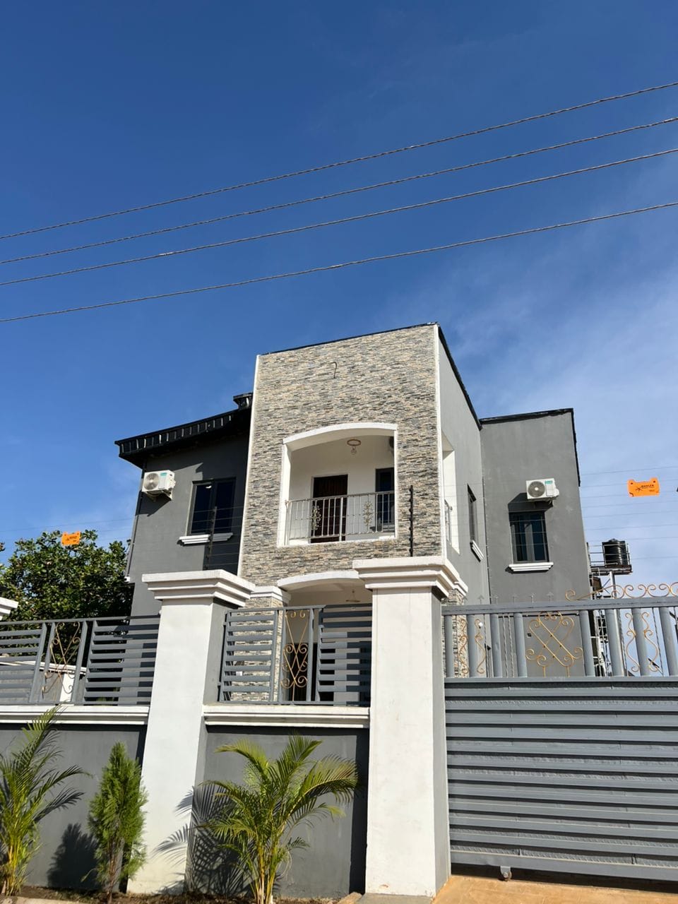 The exterior of the home features a modern design with a stone-accented entrance. A balcony is visible on the upper level, framed by a wrought iron railing. The property is enclosed by a decorative fence and includes minimal landscaping with small palm trees.
