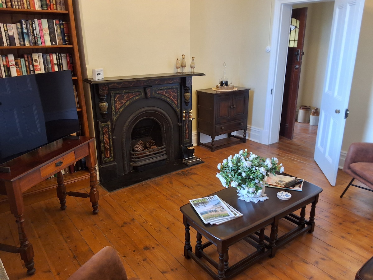 A cozy living area features wood flooring and a fireplace framed by intricate detailing. A coffee table sits adorned with a magazine and decorative items. Bookcases line one wall, and a doorway leads to another room, adding a sense of spaciousness.