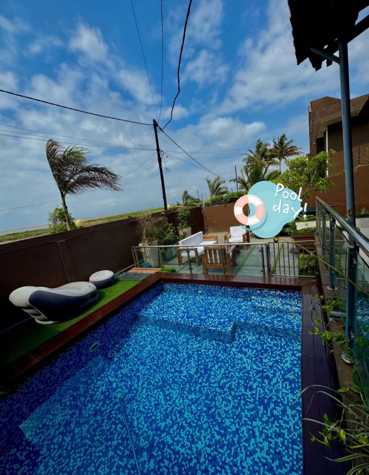 A swimming pool is surrounded by sleek glass fencing, providing an open view of the landscape. Comfortable lounge chairs sit beside the water, while lush greenery and palm trees create a peaceful backdrop under a partly cloudy sky.