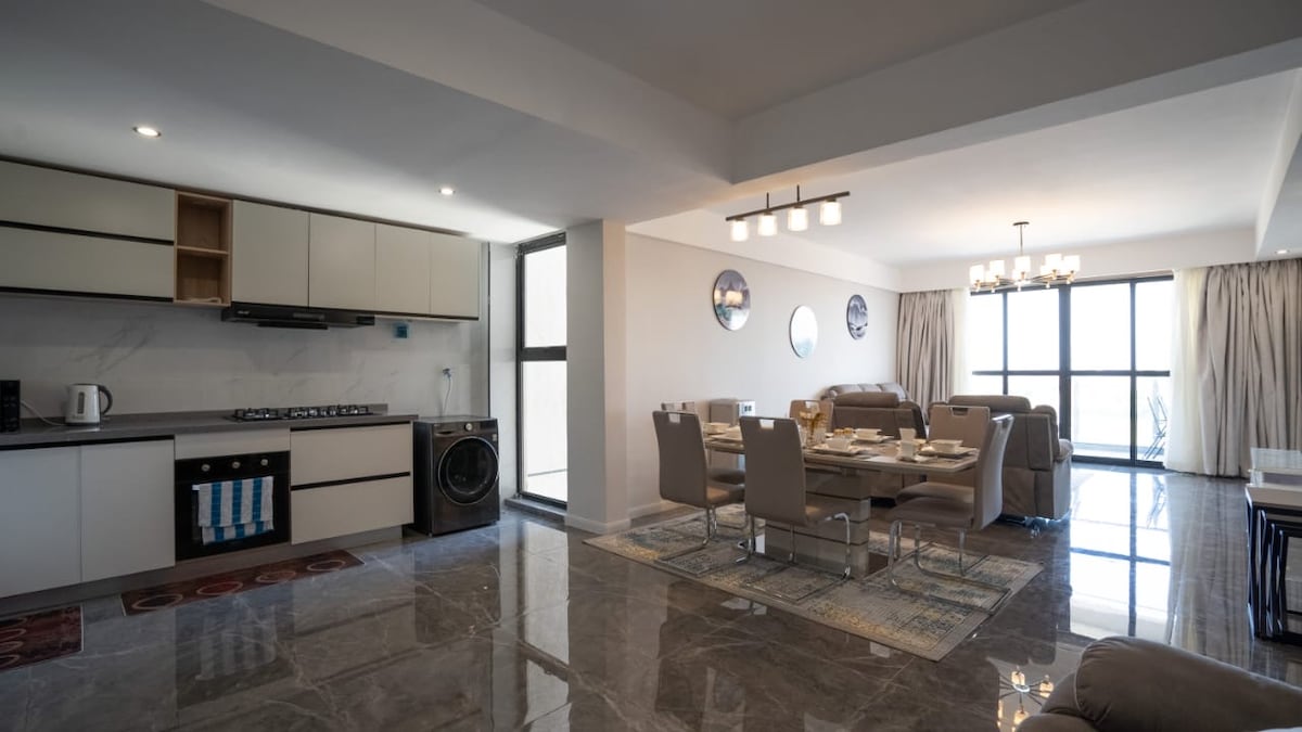 The open-plan living space showcases a modern kitchen with sleek cabinetry, a five-burner cooker, and a washing machine. A dining table for six is positioned beside large windows that provide ample natural light, enhancing the airy atmosphere of the room.