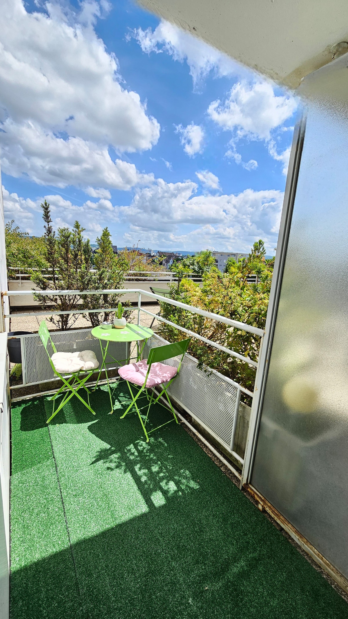 A balcony area is depicted, featuring a small round table with two chairs. One chair is green, while the other is pink. Lush greenery is visible in the background, and the bright blue sky is adorned with fluffy clouds, creating a cheerful atmosphere.