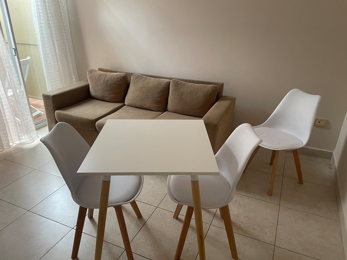 A small dining area is featured, consisting of a white table surrounded by three modern chairs. A comfortable beige sofa is positioned nearby. Natural light enters through sheer curtains, illuminating the simple and inviting space.