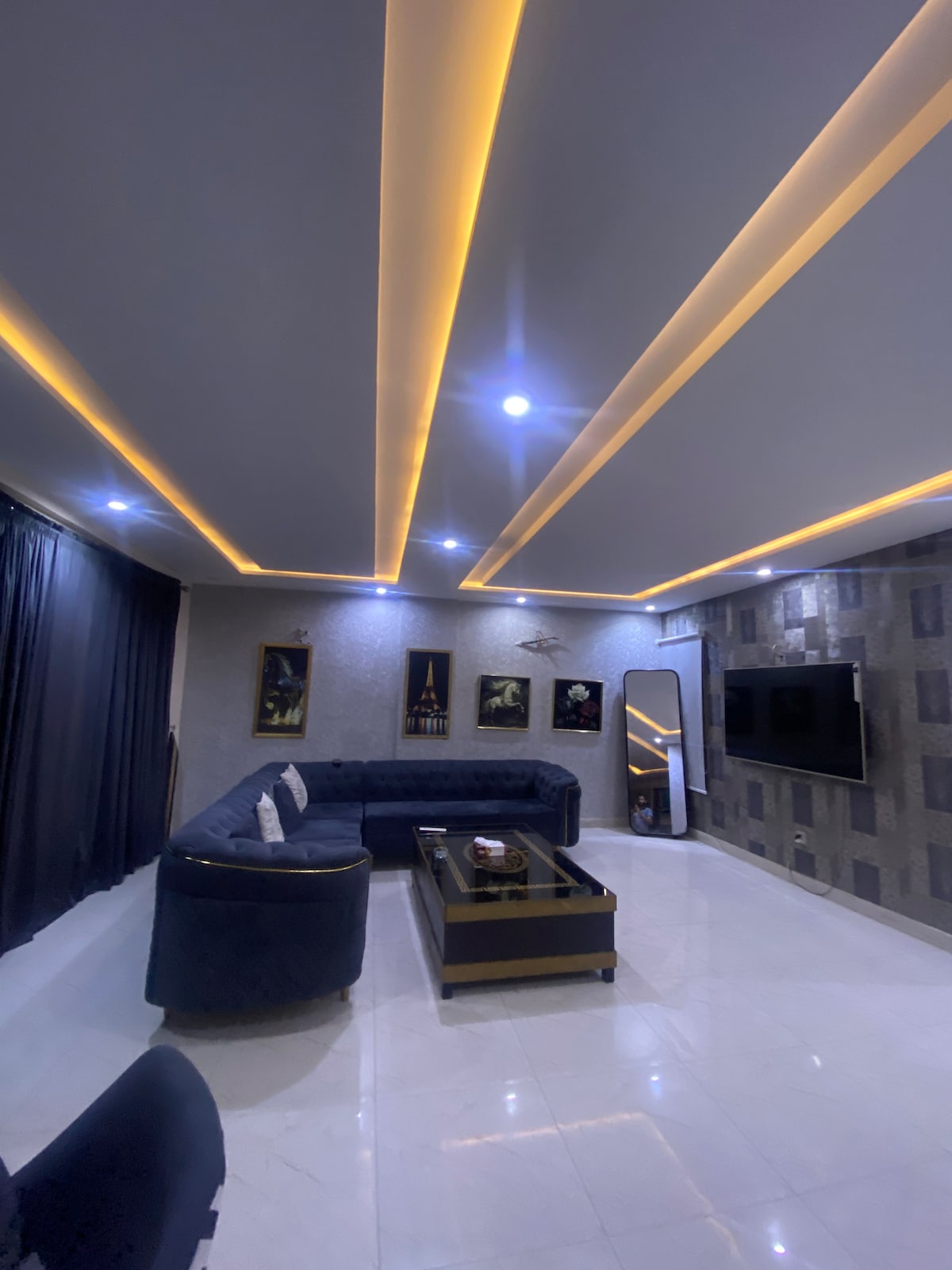 A modern living room features sleek furnishings with a dark sectional sofa, a contrasting coffee table, and a large wall-mounted TV. Ambient lighting is provided by linear ceiling fixtures, while dark curtains frame the window. Artwork is displayed on the walls, adding visual interest.