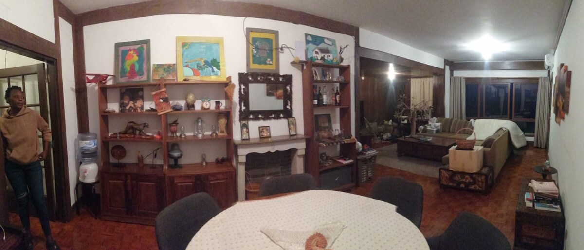 A spacious living area features a central dining table surrounded by chairs. Bookshelves line one wall, displaying decorative items and artwork. A comfortable seating arrangement is visible in the background, complemented by large windows allowing natural light to fill the space.