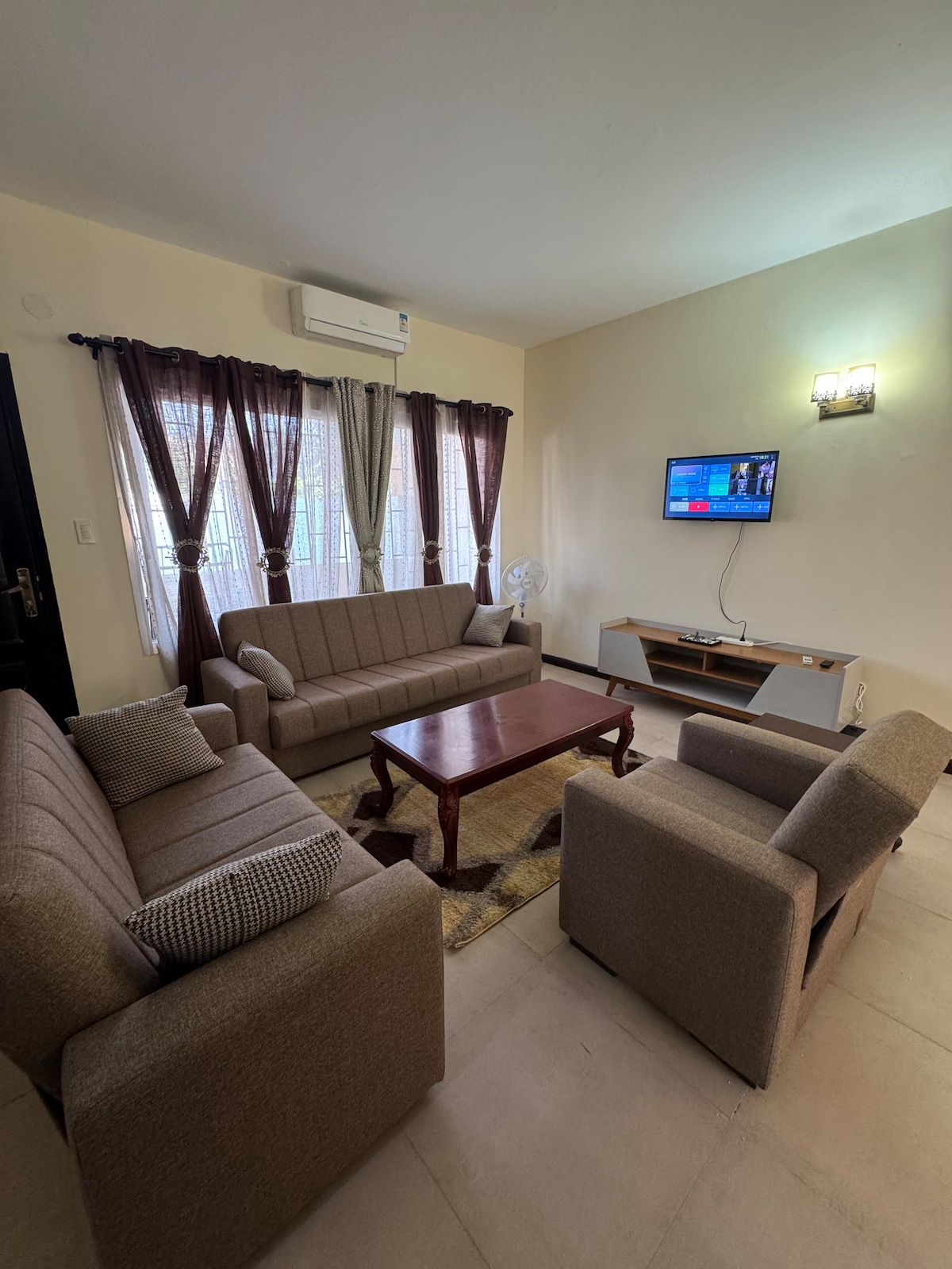 A welcoming living area is furnished with multiple sofas in neutral tones, surrounding a wooden coffee table. Large windows are dressed with sheer curtains, allowing natural light to filter in. A television is mounted on the wall opposite the seating arrangement.