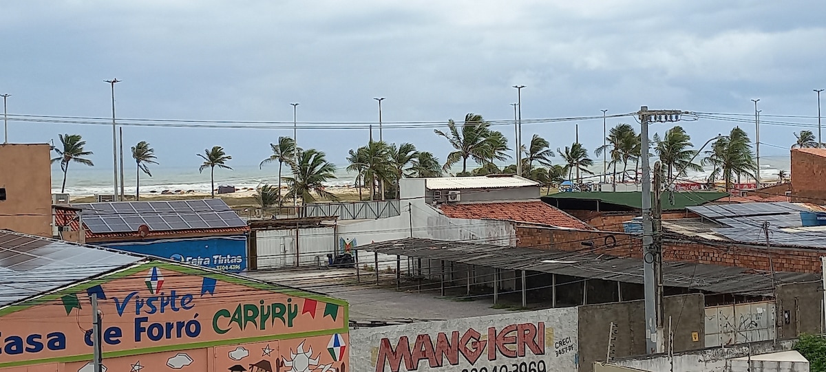 An expansive view showcases a vibrant beachside locale, with palm trees swaying along the shoreline. Colorful storefronts, including a prominent house of forró, add character to the scene, while the ocean is visible in the distance beneath a cloudy sky.