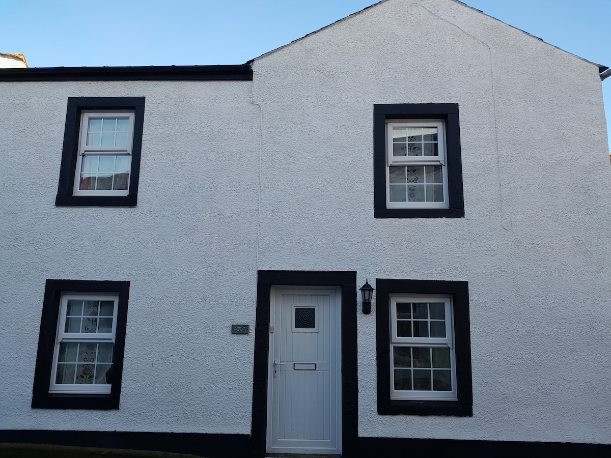 The exterior of Ashton Cottage features a white facade with black trim. The entrance includes a white door, flanked by three windows with decorative panes. Natural light reflects off the exterior, enhancing the charming character of this converted barn.