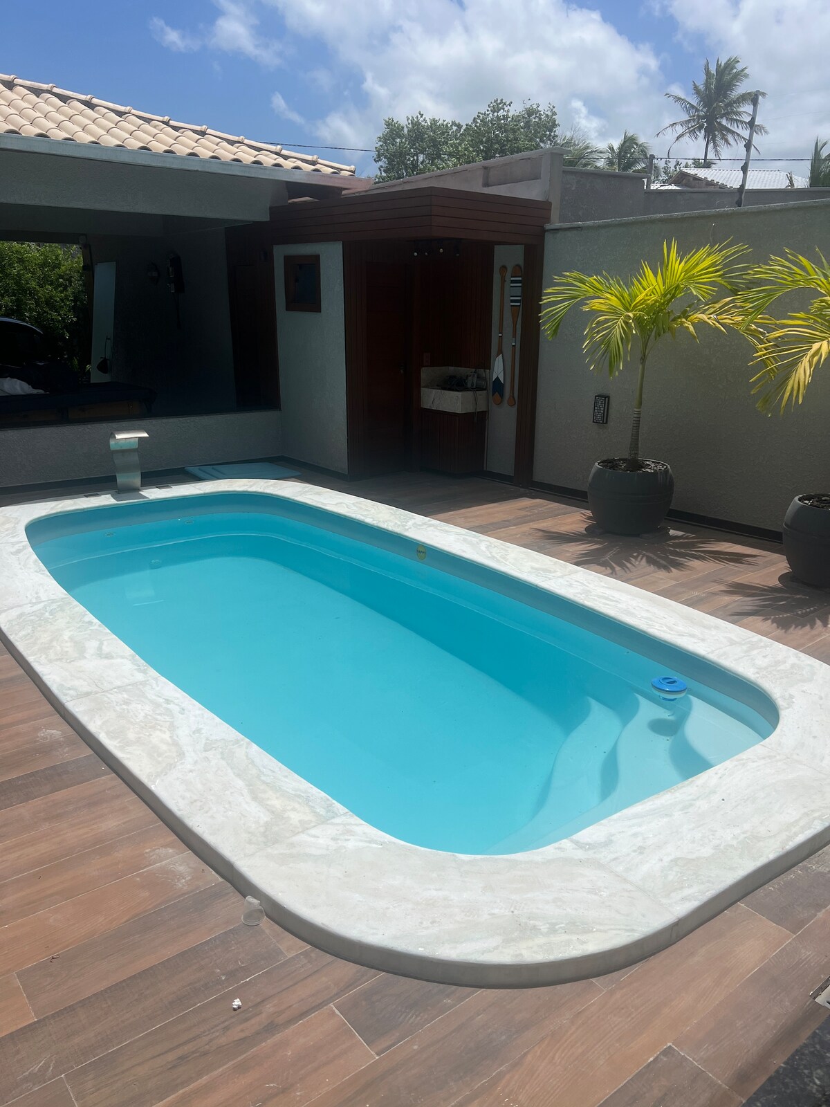 A clear, inviting pool is surrounded by natural wood decking. Lush greenery is visible in the background, complementing the serene atmosphere. Sunlight reflects off the water, creating a refreshing space for relaxation.