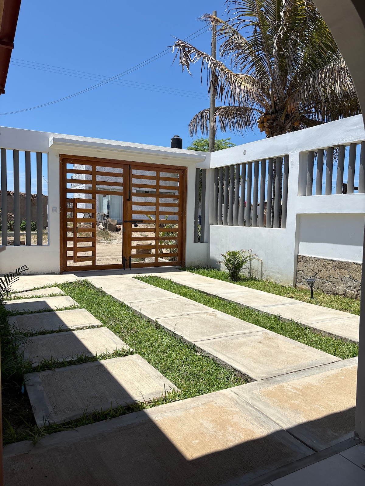 A welcoming entrance features a wooden gate with horizontal slats, surrounded by a neatly landscaped yard. Concrete stepping stones create a pathway through lush green grass, leading toward a bright blue sky. Tall palm trees are positioned nearby, adding to the tropical ambiance.