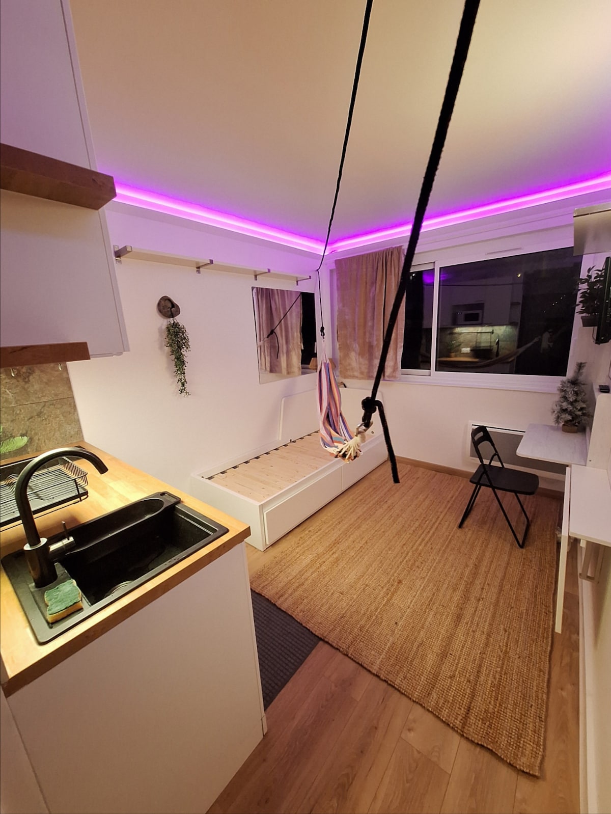 A cozy studio space features a swing hammock suspended from the ceiling, alongside a minimalist kitchen area with a sleek black sink. A foldable chair and desk are positioned near a window with curtains, illuminated by soft, adjustable LED lighting along the ceiling.