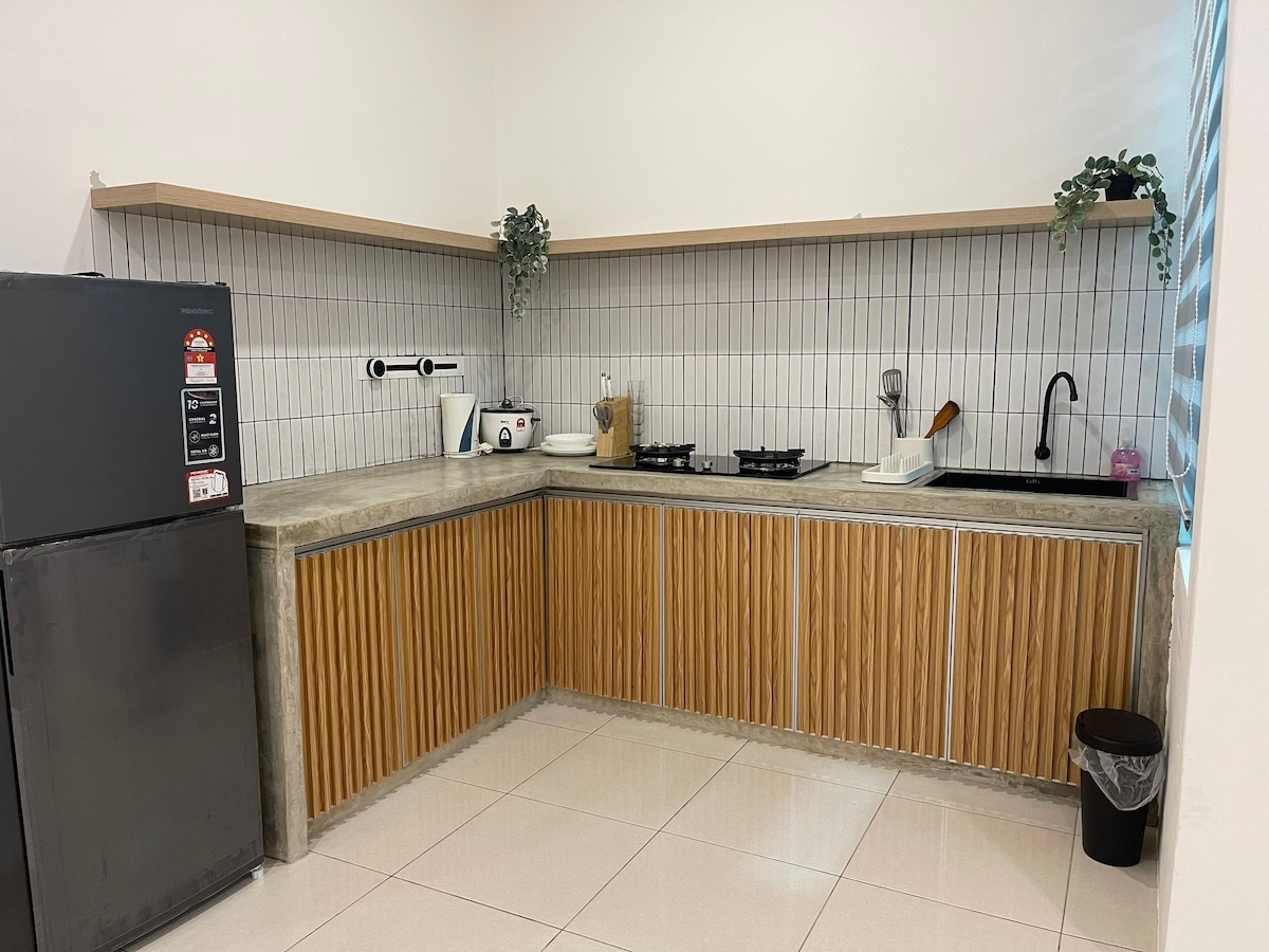 A modern kitchen features sleek concrete countertops and wooden cabinetry. A refrigerator stands to the left, with small appliances arranged on the counter. The cooking area is equipped with a gas stovetop and a sink, complemented by minimalistic decor and potted plants.