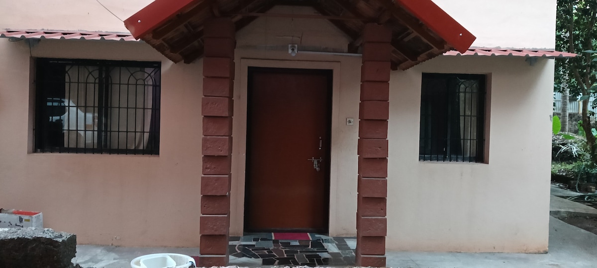 The entrance features a wooden door framed by two stone columns, with a covered porch above. Symmetrical windows with metal grilles are present on both sides. A stone pathway leads to the door, and a small outdoor area is visible in front.