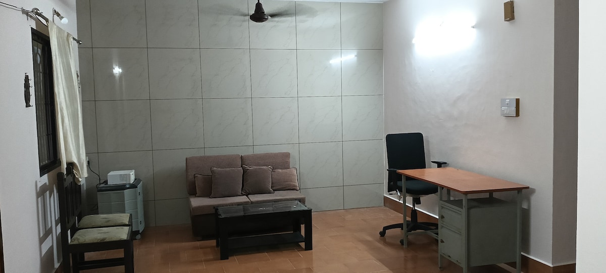 A spacious living area features a comfortable sofa set, a coffee table, and a desk with an ergonomic chair. Soft lighting is provided by wall-mounted fixtures, and the floor is finished in warm tiles. A light-colored curtain frames the window, enhancing the relaxed atmosphere.
