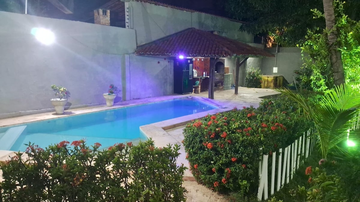 A tranquil outdoor space is illuminated at night, featuring a clear swimming pool surrounded by lush greenery and colorful flowers. A traditional roofed area is visible nearby, adding a rustic charm to the setting.