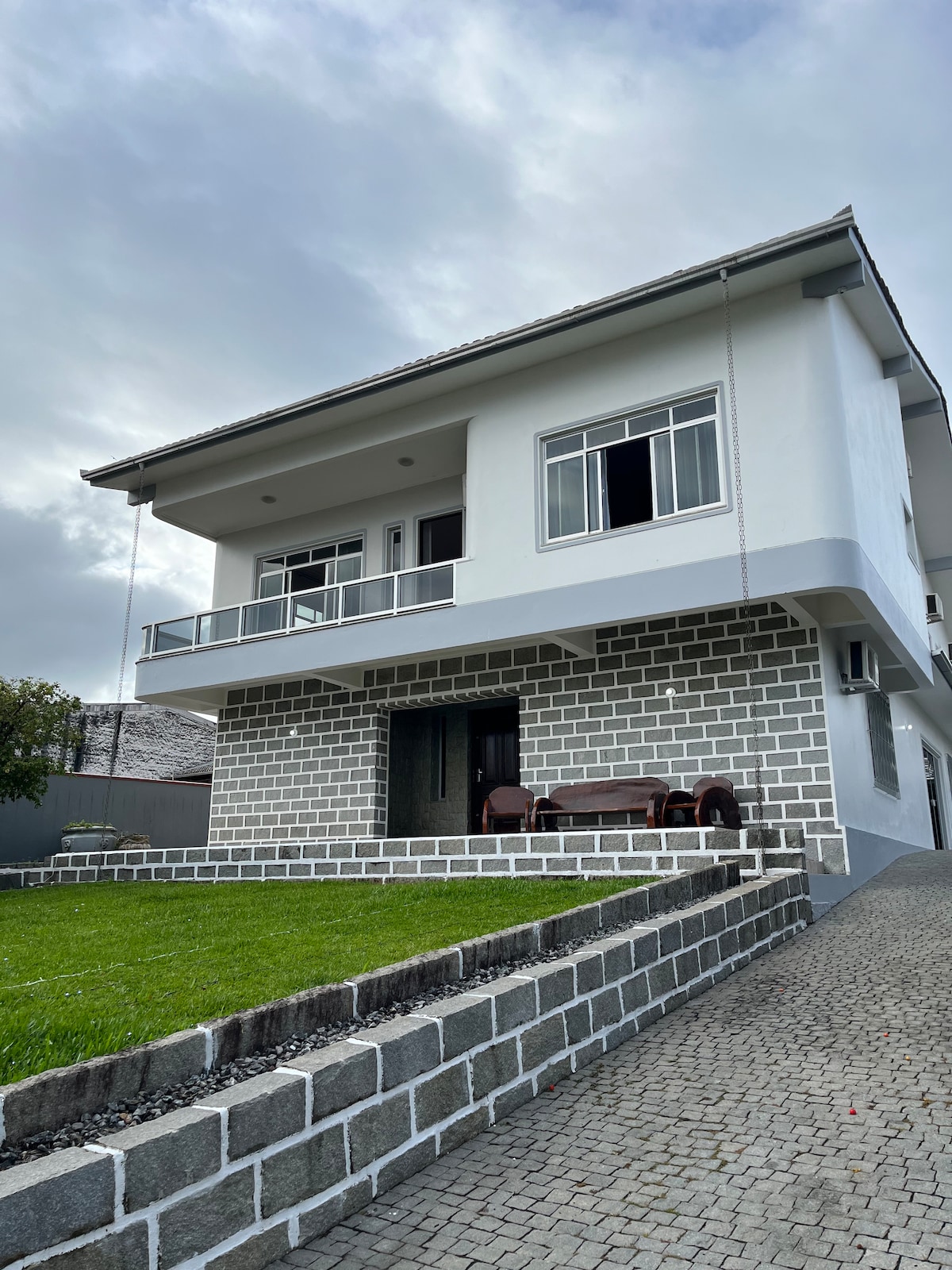 A two-story house is depicted with a modern exterior featuring a light-colored facade and a stone base. A balcony extends over the front, and large windows provide natural light. A stone pathway leads to an inviting front porch with a seating area, surrounded by a manicured lawn.