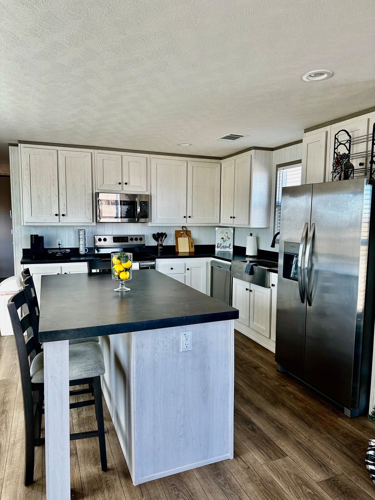 A modern kitchen is presented with sleek white cabinetry and a dark countertop. Stainless steel appliances include a refrigerator and microwave. A central island with bar seating displays a bowl of fresh fruit, contributing to the functional and inviting layout.