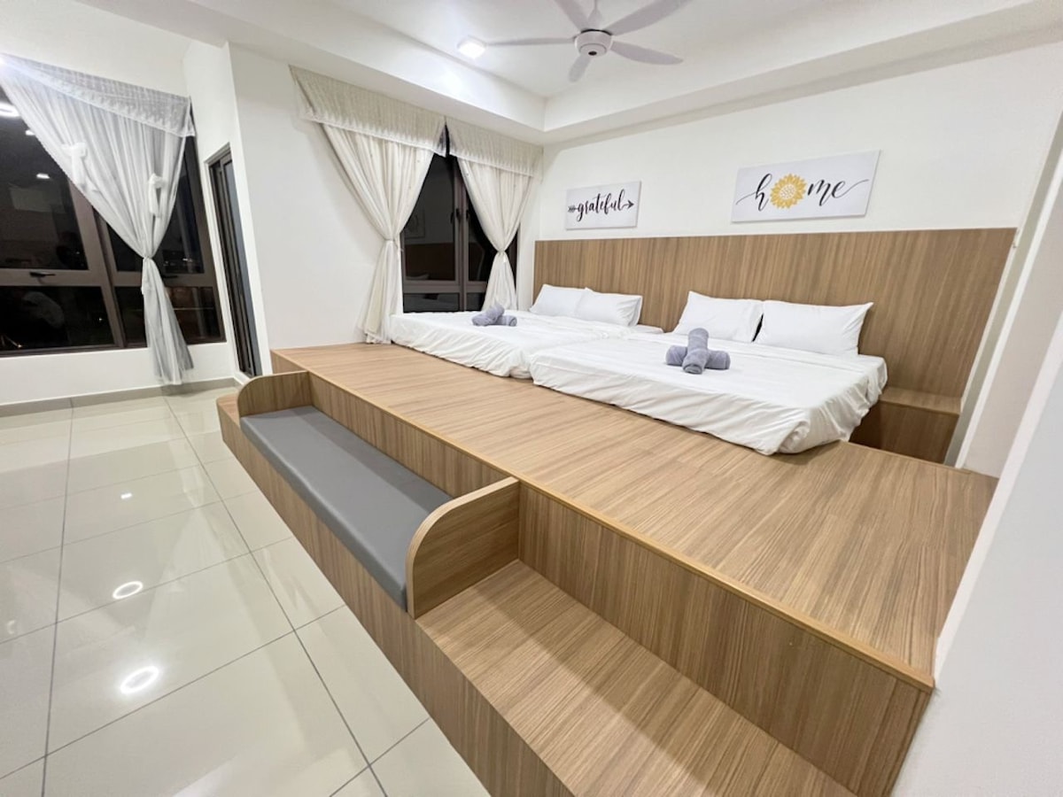 A spacious sleeping area is highlighted by two beds set on a raised platform, featuring simple white linens and towels arranged neatly. Large windows allow natural light to fill the room, complemented by sheer curtains. Decorative wall art enhances the modern design.