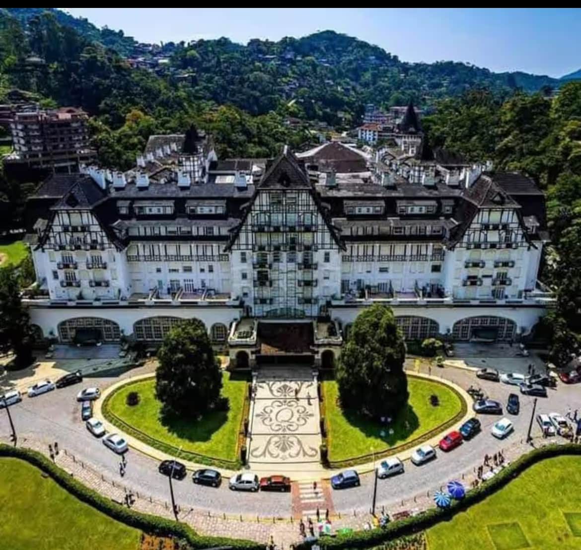 An aerial view of a large, historic building surrounded by lush greenery and landscaping. The structure features distinct architectural elements, including gabled roofs and numerous windows. Vehicles are parked along a circular driveway, enhancing the sense of community and accessibility.