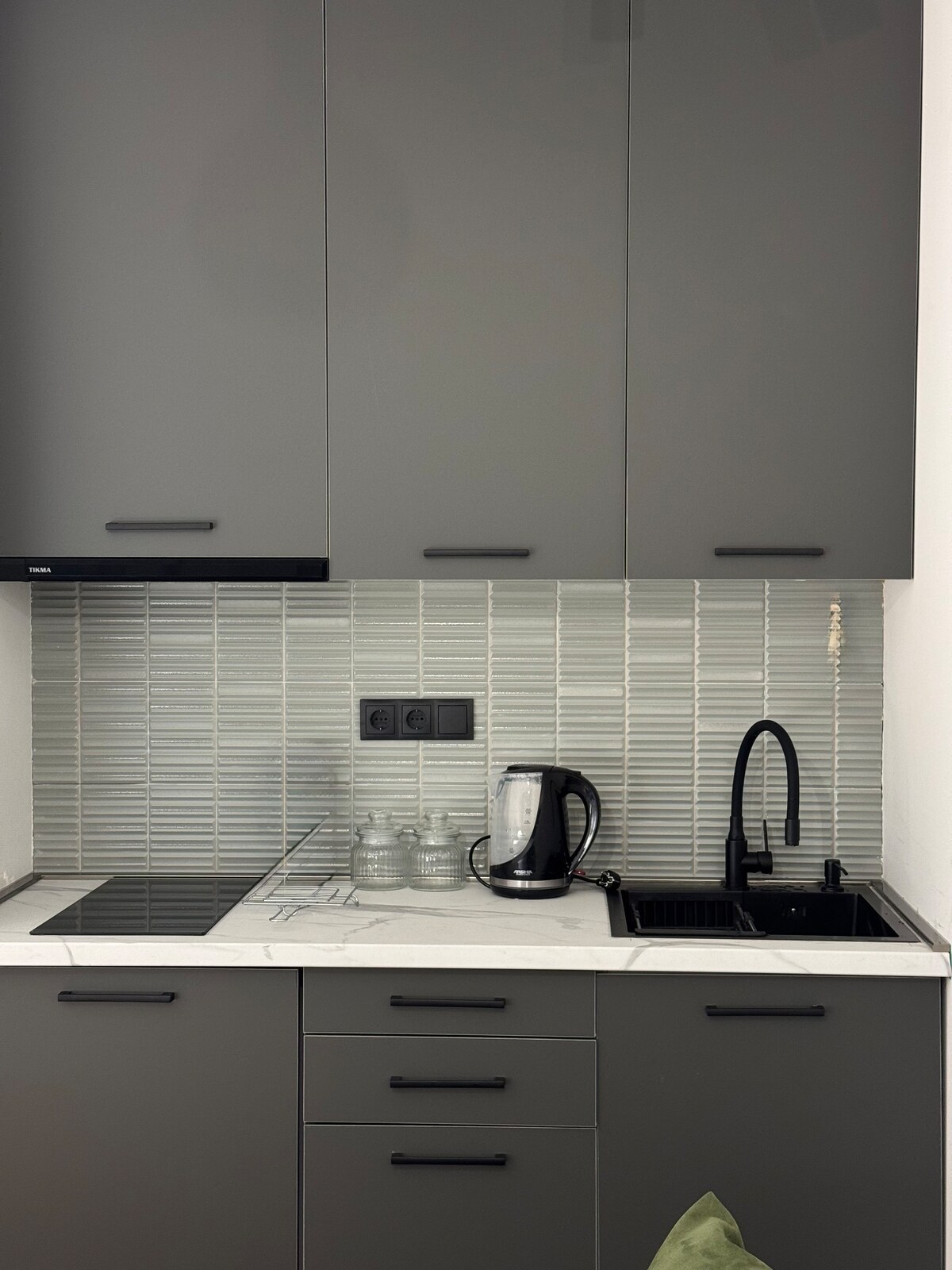 A modern kitchen area is fitted with sleek grey cabinetry and a stylish backsplash. An electric kettle and a glass container are placed on the countertop. A black sink is integrated alongside an electric cooktop, providing a convenient cooking space.