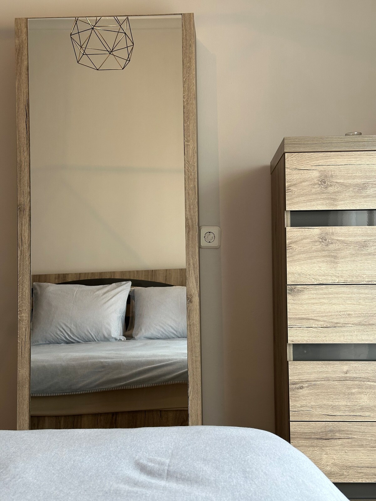 A full-length mirror set against a pale wall reflects a cozy sleeping area with a soft gray blanket draped over the spacious bed. A modern wooden dresser with multiple drawers stands beside the mirror, complementing the overall design.