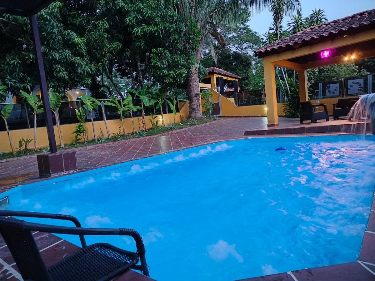 A tranquil pool with clear blue water is set within lush greenery and palm trees. A shaded area is provided nearby, featuring seating and a cascading waterfall that flows gracefully into the pool.