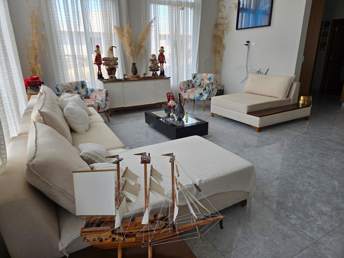 A modern living area features a spacious beige sofa adorned with soft pillows. A low coffee table sits in the center, accompanied by decorative items. Light floods the room through large windows, illuminating a nearby unique sculpture on a white cabinet.