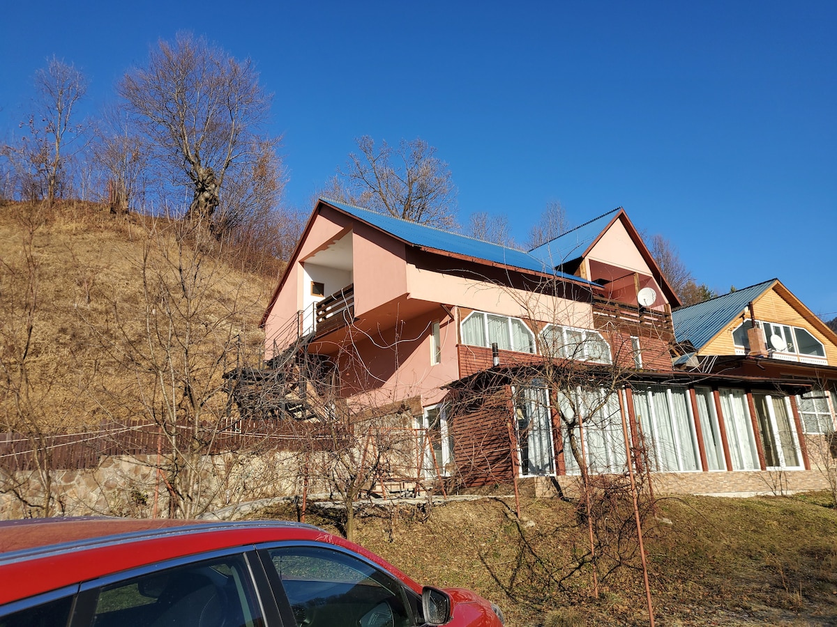 A multi-level building sits against a clear blue sky, featuring large windows that reflect natural light. The structure is complemented by an outdoor terrace, with a sloping hillside and sparse vegetation visible in the foreground.