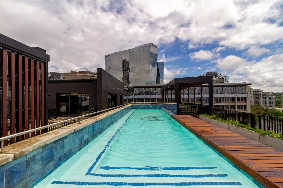 A rooftop pool is seen featuring clear turquoise water and a wooden deck. Surrounding buildings and a sculpture are visible in the background, creating a dynamic urban skyline.
