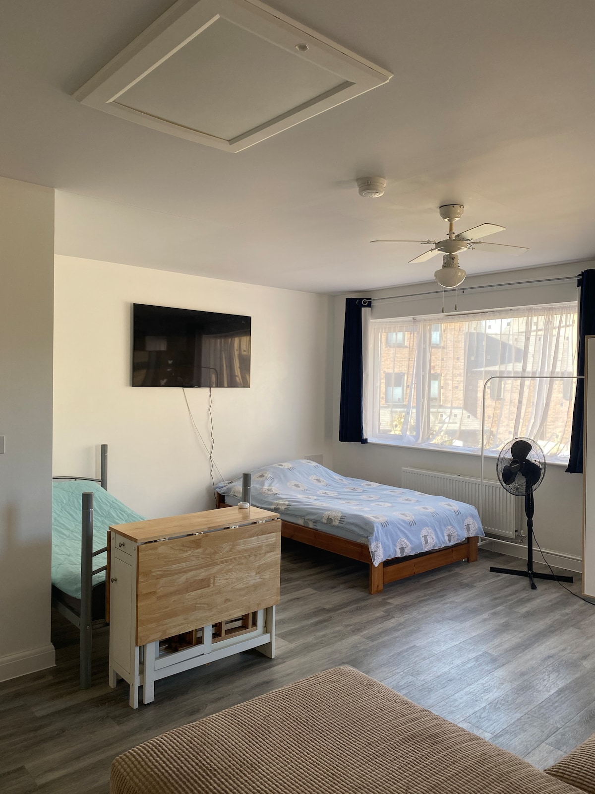 A bright studio space features a small double bed with light bedding and a single bed with a patterned comforter. A flat-screen TV is mounted on the wall. Natural light enters through large windows adorned with dark curtains, and a fan is positioned nearby.