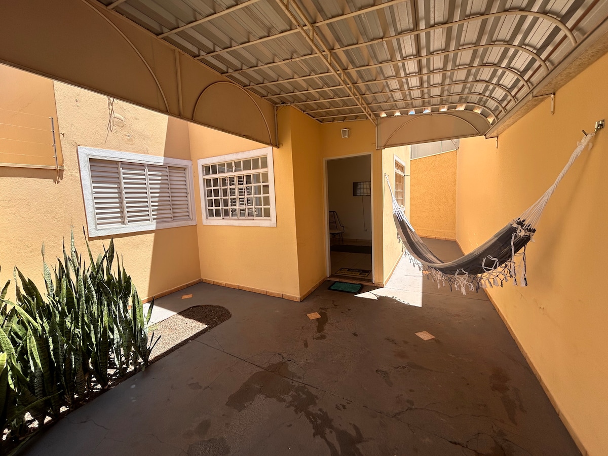 An outdoor area features a covered space with a hammock hanging between two posts. A smooth concrete floor is visible, accompanied by a small garden with green plants along the left side. The walls are painted a warm yellow, enhancing the inviting environment.