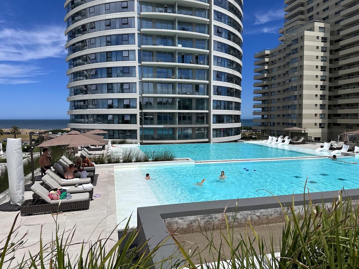 The exterior pool area is visible, featuring a large, inviting swimming pool surrounded by comfortable lounge chairs and umbrellas. The modern building reflects a coastal backdrop, with the ocean visible in the distance. A well-maintained landscape accompanies the serene atmosphere.