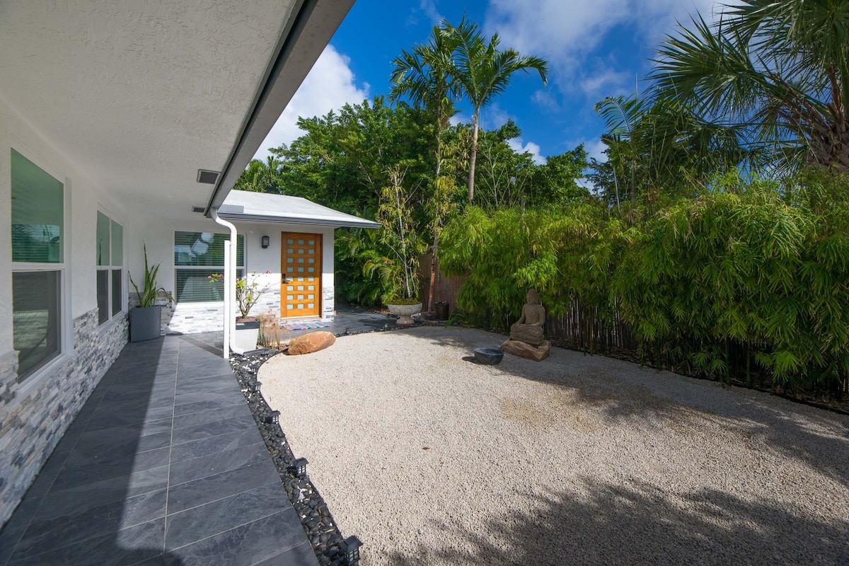 A serene outdoor area features a gravel pathway leading to a modern entrance with a vibrant orange door. Lush greenery, including palm and bamboo plants, creates a natural barrier, while a stone sculpture is subtly placed in the landscape.