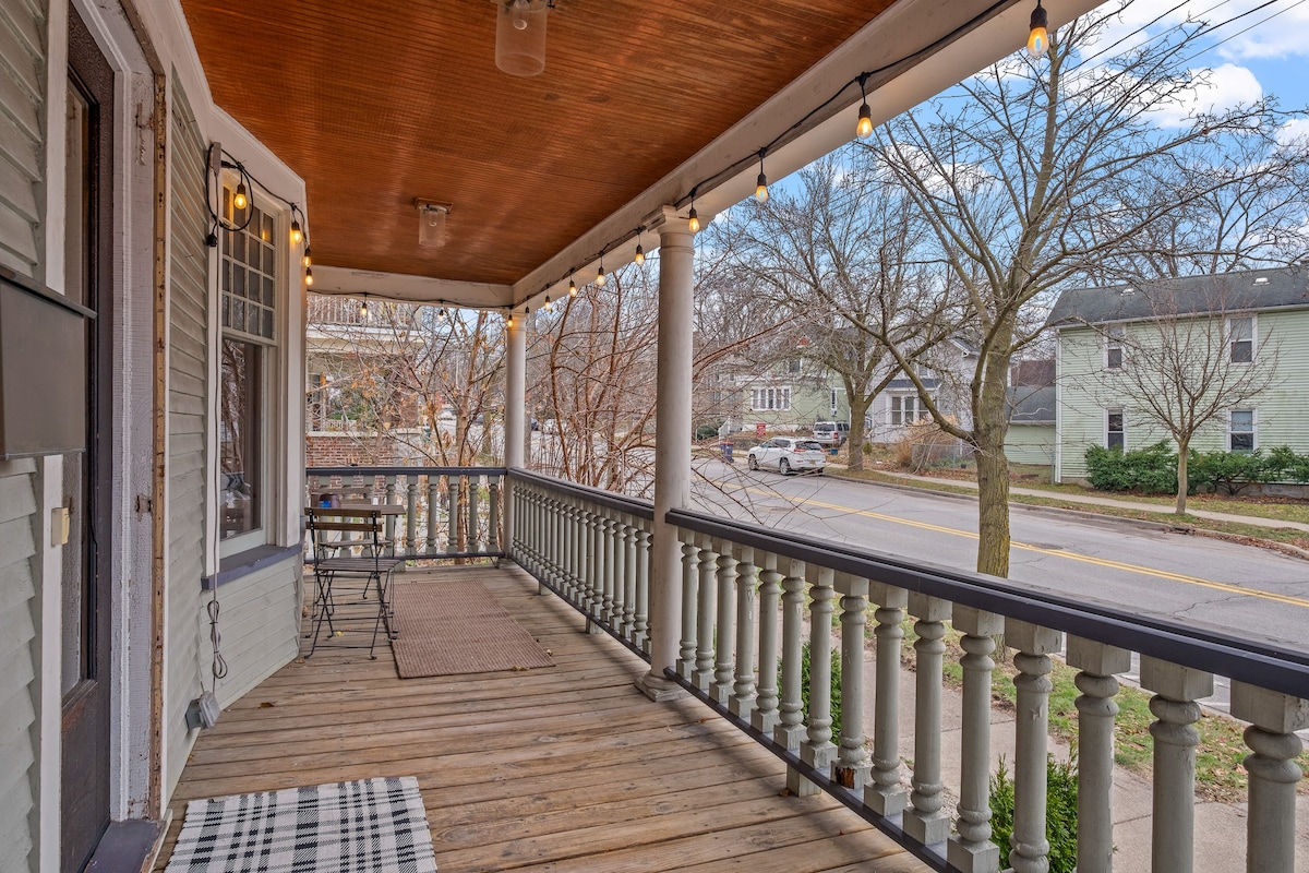 A covered porch with wooden flooring is outlined by decorative railings. String lights hang above, enhancing evening ambiance. A small table and chairs are positioned on one side, while the street view includes trees and neighboring houses. The space is inviting for outdoor relaxation.